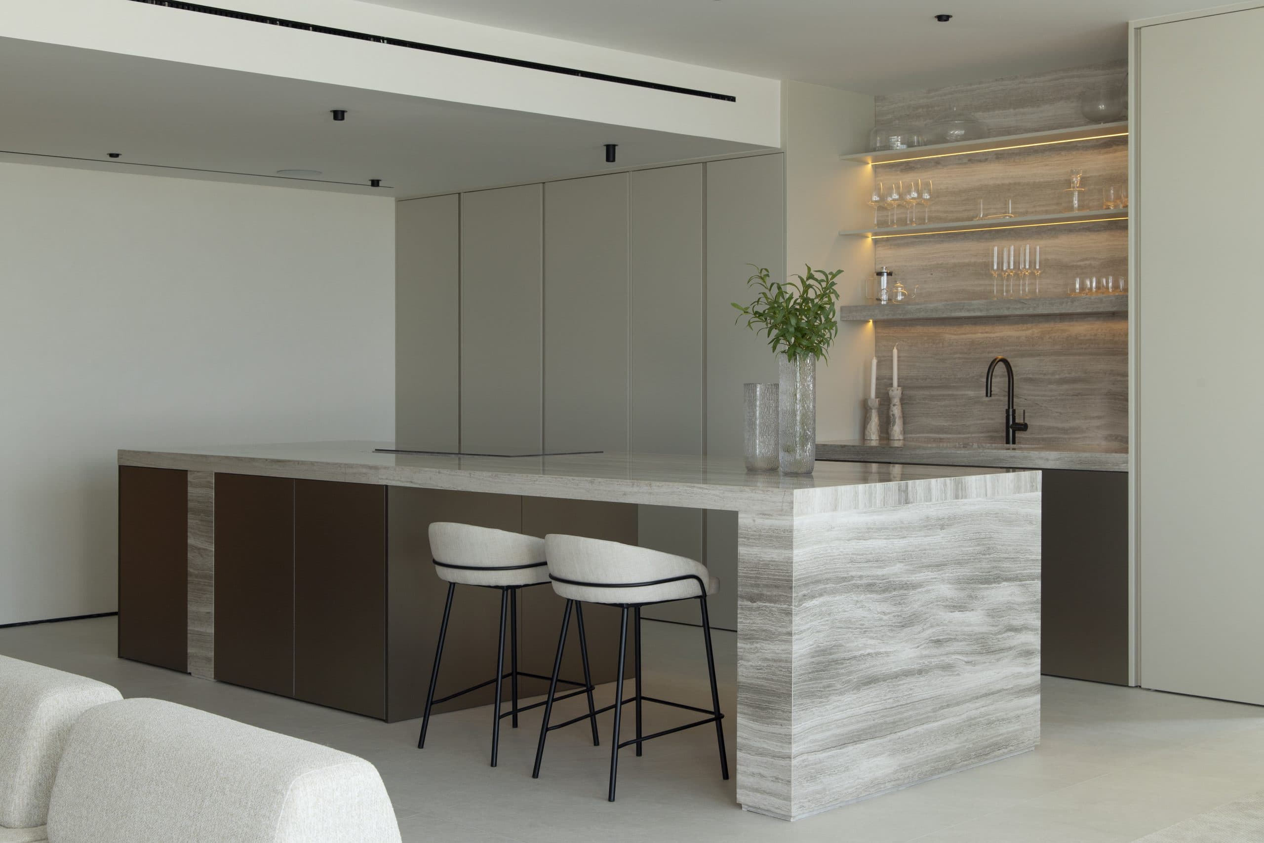 Kitchen island clad in veined stone with dark oak base units and lit glass shelving behind